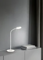 De TRIO Tafellamp Monza, een witte bureaulamp met een flexibele hals, staat op een grijze kast naast drie gestapelde notitieblokken. De lamp brandt en werpt licht op de kast, met een grijze muur en donkere gordijnen op de achtergrond.