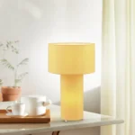 De TRIO Tafellamp Bale in geel staat op een wit oppervlak naast een houten dienblad met een witte theepot en beker, terwijl potplanten en lichte gordijnen een zacht verlichte achtergrond creëren.