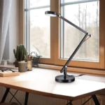 De TRIO Tafellamp AMSTERDAM, een strakke zwarte bureaulamp, staat op een houten bureau bij een raam naast een cactus in pot, een notitieblok en een kopje, met natuurlijk licht dat naar binnen stroomt.