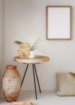 In een minimalistische kamer staat de REALITY Tafellamp JEAN op een ronde houten tafel met zwarte poten, een geweven mand, een kleine witte vaas met gedroogd gras, een grote keramische vaas met structuur op de vloer, een beige kussen en een leeg houten fotolijstje aan de muur.