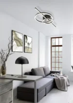 Moderne woonkamer met een grijze bank, grijze deken, abstracte muurkunst, bureau met de TRIO Tafellamp Nola en een unieke ronde plafondlamp. Het grote raam vult de ruimte met natuurlijk licht.
