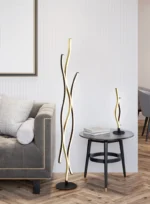 Een moderne woonkamer toont een grijze getufte sofa met een kussen met patroon, een zwarte bijzettafel met daarop de TRIO Tafellamp Blaze golvende LED lamp en een bijpassende hoge vloerlamp met golvende LED lichtstrips.