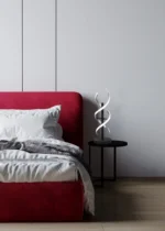 Een moderne slaapkamer met een rood gestoffeerd bed, wit beddengoed en een zwart nachtkastje met de TRIO Sequence tafellamp, tegen een effen witte muur en een lichthouten vloer.