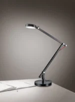 De TRIO Tafellamp Amsterdam, een moderne zwarte bureaulamp met een verstelbare arm, is verlicht en geplaatst boven een open notitieblok op een wit bureau tegen een eenvoudige grijsverloopachtergrond.
