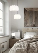 Een gezellige slaapkamer heeft een beige gebreide deken op het bed, een houten hoofdeinde, een grote rustieke spiegel en een nachtkastje met de REALITY Tafellamp MADELINE. Twee moderne hanglampen hangen erboven, terwijl natuurlijk licht door het raam naar binnen stroomt.