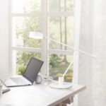 In een zonovergoten kamer staat de TRIO Tafellamp Manduro op een wit bureau met een open laptop, witte koptelefoons, papieren en ramen met transparante gordijnen die groene bomen buiten laten zien.