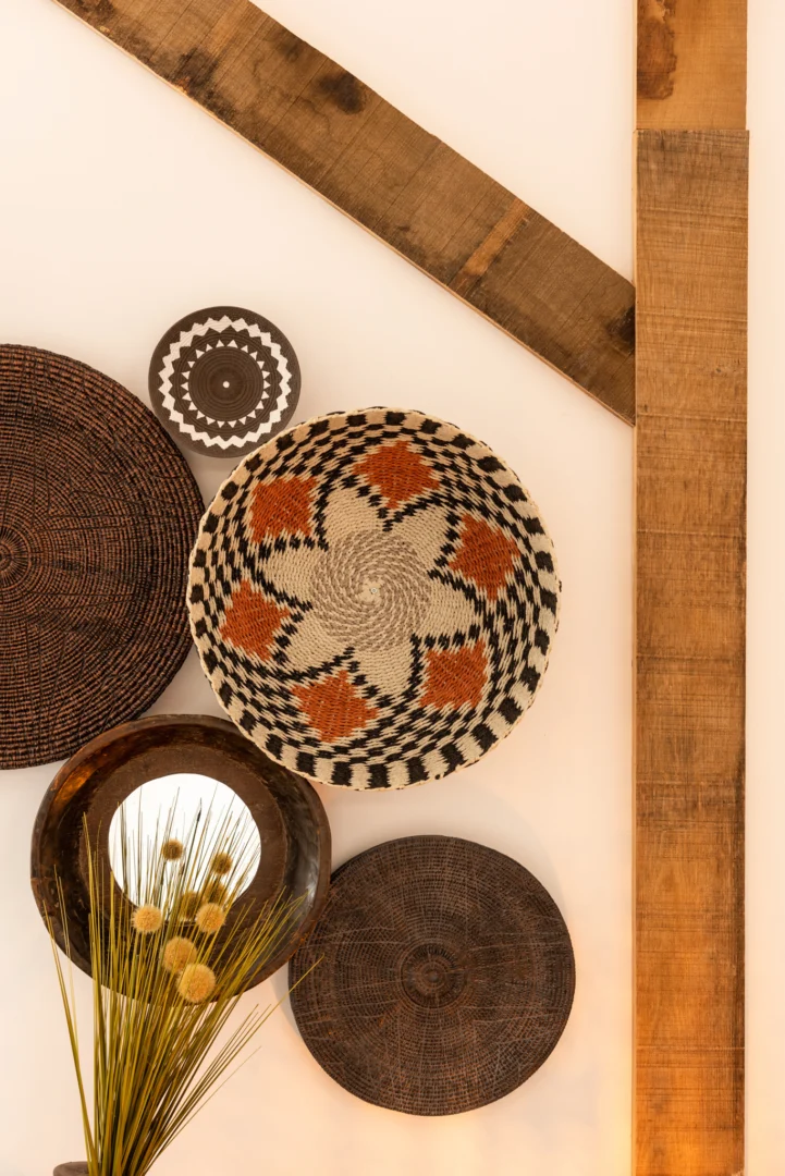 Aan de muur hangt de Mirror Round Bowl Shaped Recycled Wood Brown, gecombineerd met gevlochten manden in zwart, bruin, wit en oranje patronen, houten planken en een vaas met gedroogde gele bloemen.