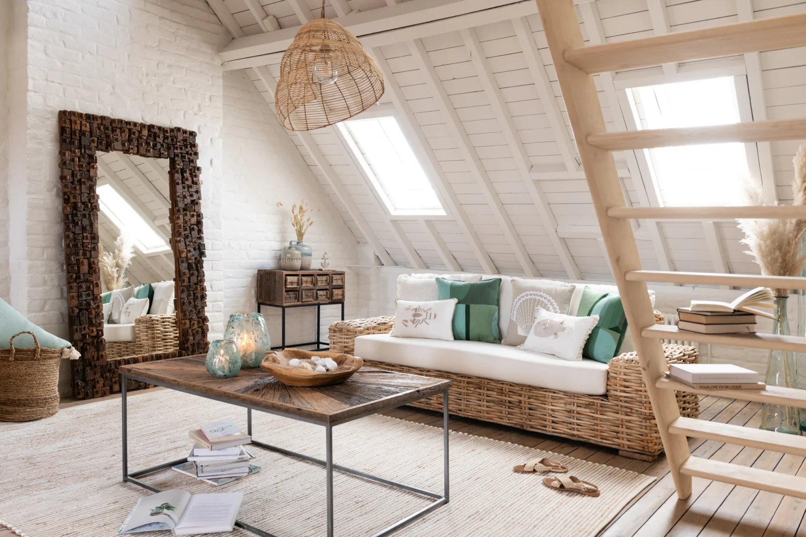 Een lichte zolderkamer heeft een geweven sofa, groene en witte kussens, een rustieke salontafel en de MIRROR SYMA RECYCLED WOOD/MIRROR NATURAL LARGE, met natuurlijk licht dat door dakramen in het plafond met witte lambrisering naar binnen stroomt.
