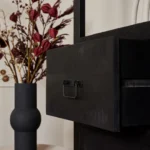 Een close-up van een open zwarte Boekenkast Boaz Black | 3 laden lade met een metalen handvat, naast een matzwarte vaas met gedroogde rode en bruine bloemen, tegen een zacht verlichte neutrale achtergrond.