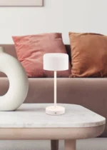 De REALITY Tafellamp JEFF, een moderne witte tafellamp, staat op een marmeren salontafel voor een beige bank met roze en roestkleurige kussens, naast een witte sculpturale vaas.