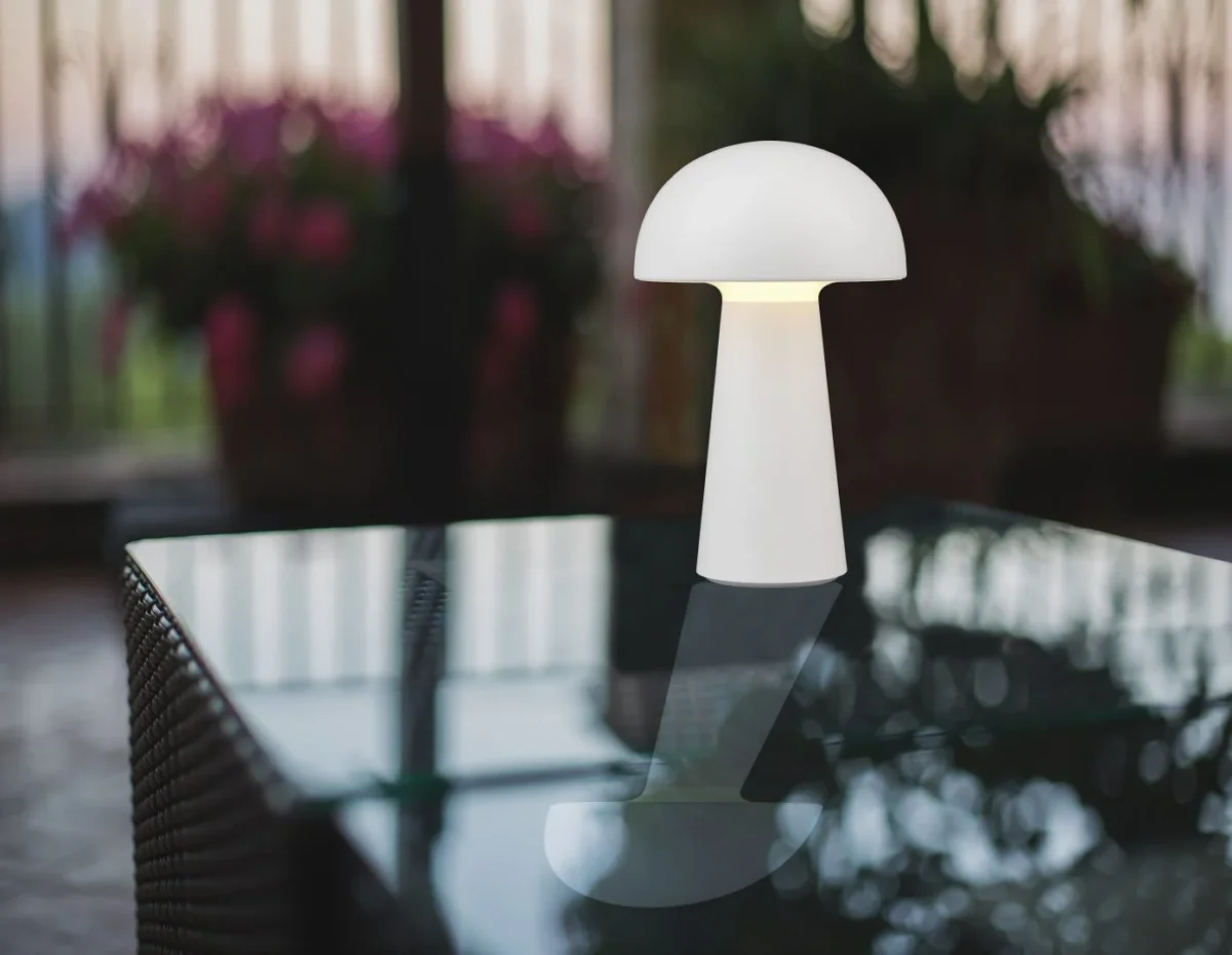 De REALITY Tafellamp LENNON, een moderne witte paddenstoelvormige lamp, staat op een glazen buitentafel met vage potplanten en balustrades op de achtergrond.