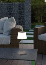De Reality Tafellamp Sanchez, een kleine tafellamp met een witte kap, staat op een stenen buitentafel temidden van rieten terrasmeubilair met lichte kussens. Groene bomen en een stenen structuur staan op de achtergrond.