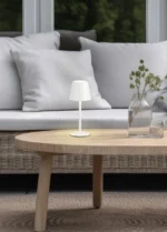 De Reality Tafellamp Diaz, een kleine witte tafellamp, staat op een lichthouten salontafel naast glazen vazen met gedroogde bloemen. Op de achtergrond staan een witgeklede bank en grote ramen met uitzicht op groen.