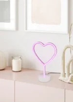 De REALITY Tafellamp HEART, een hartvormige roze neonlamp, staat op een lichtroze kast naast een witte kaars en decoratieve voorwerpen, met daarachter abstracte kunst op de witte muur.