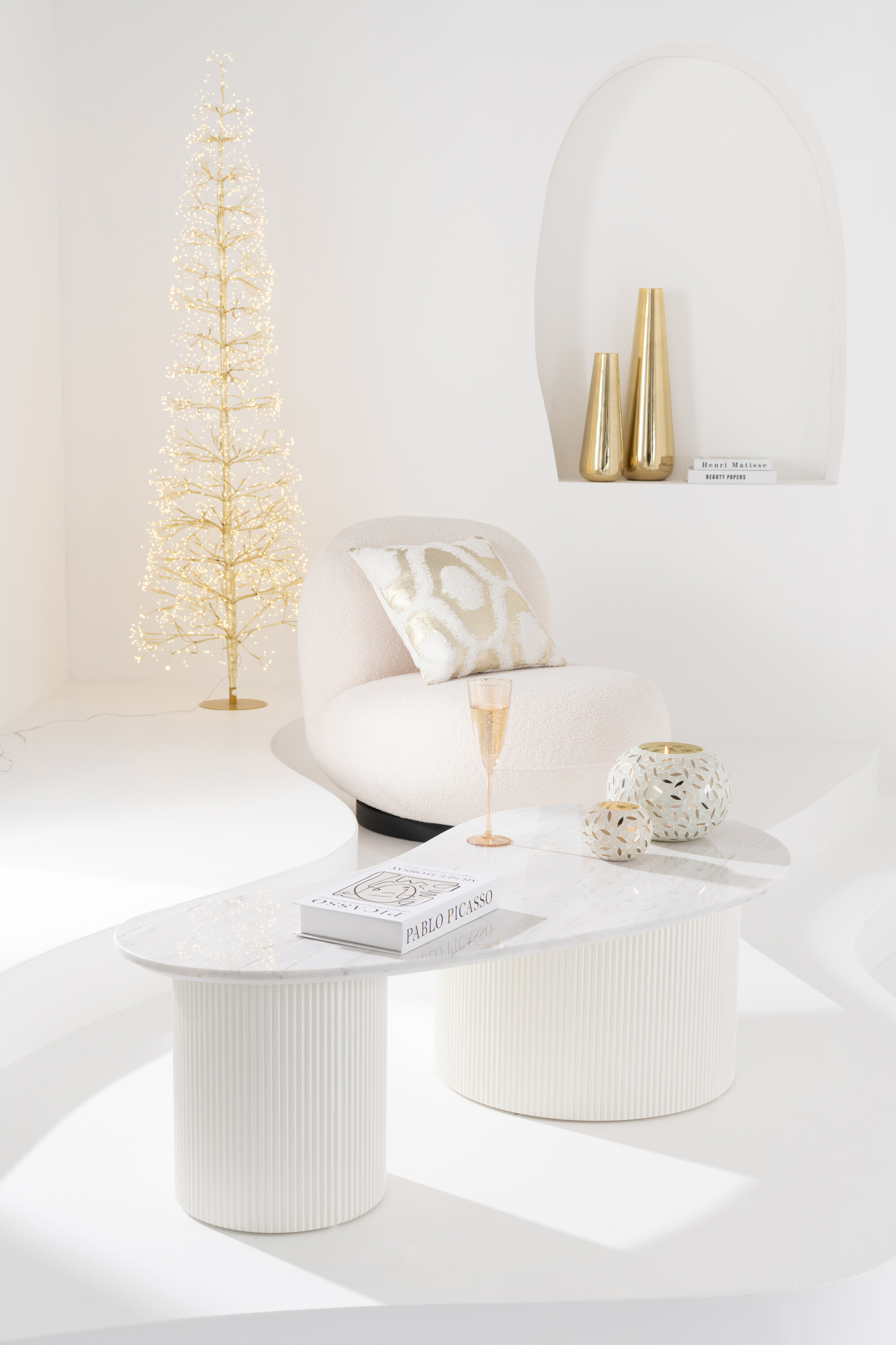 Een moderne, minimalistische woonkamer met witte muren is voorzien van de Lounge Chair Sneeuw Wit/Zwart Comfort, gouden vazen, een champagneglas, een boek, een kaars, een goudverlichte boom - allemaal gerangschikt op strakke witte gebogen tafels.