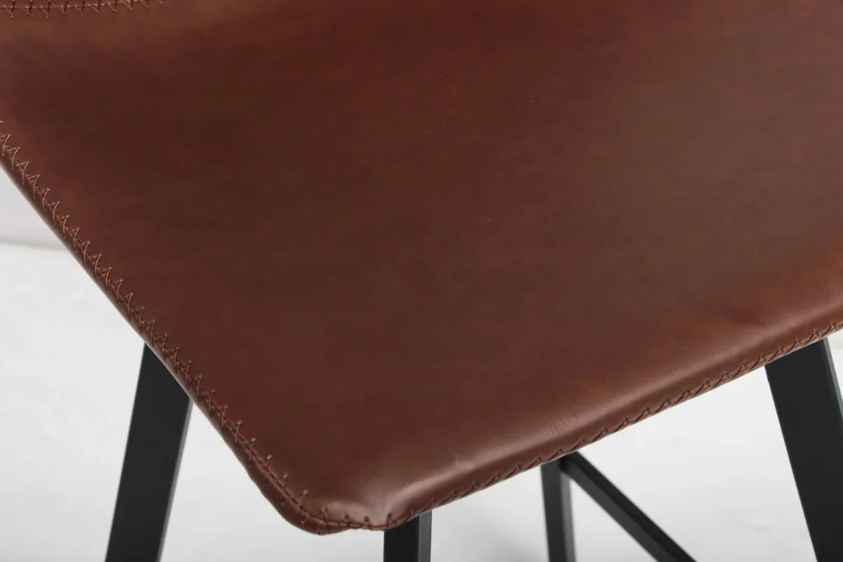 Close-up van Barstoel 'Ozan' PU Bruin zitting met zichtbare stiksels op bruin leer en gedeeltelijk zichtbare zwarte metalen poten (set van 2 per doos).