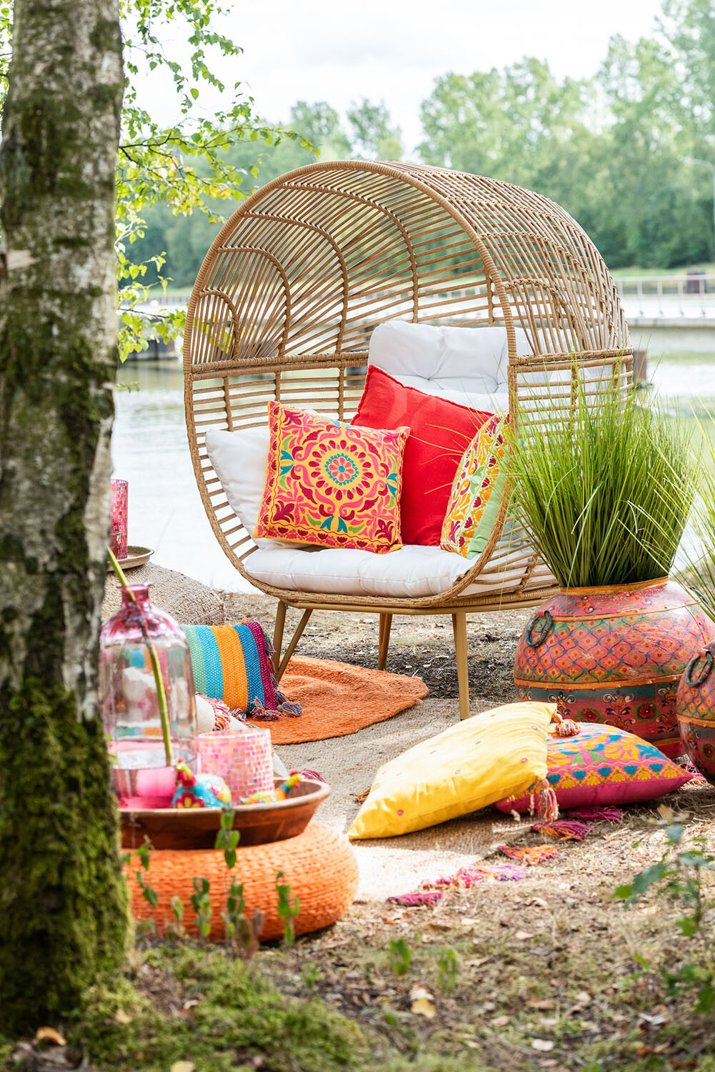 Een gezellige buitenruimte aan het meer is voorzien van de Lounge Stoel Ovaal Natuurlijk Staal met kleurrijke kussens, levendige kussens, gedessineerde tapijten, grote decoratieve vazen en wordt omringd door weelderig groen en bomen.