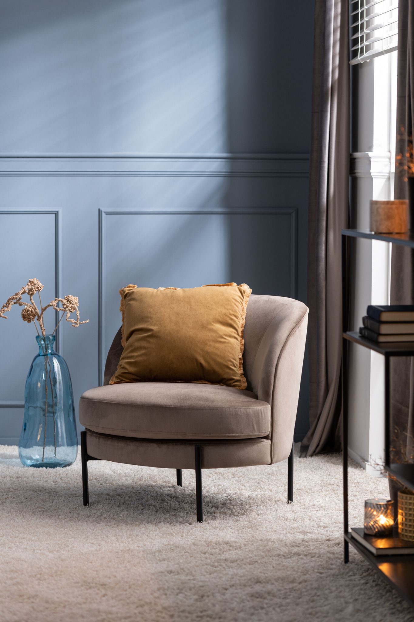 De Lounge Stoel Rond Bruin Textiel staat op een licht tapijt naast een hoge blauwe vaas gevuld met gedroogde bloemen. Zacht licht stroomt naar binnen door de transparante gordijnen, waardoor de gezellige bruine stoffen bekleding van de stoel goed uitkomt.