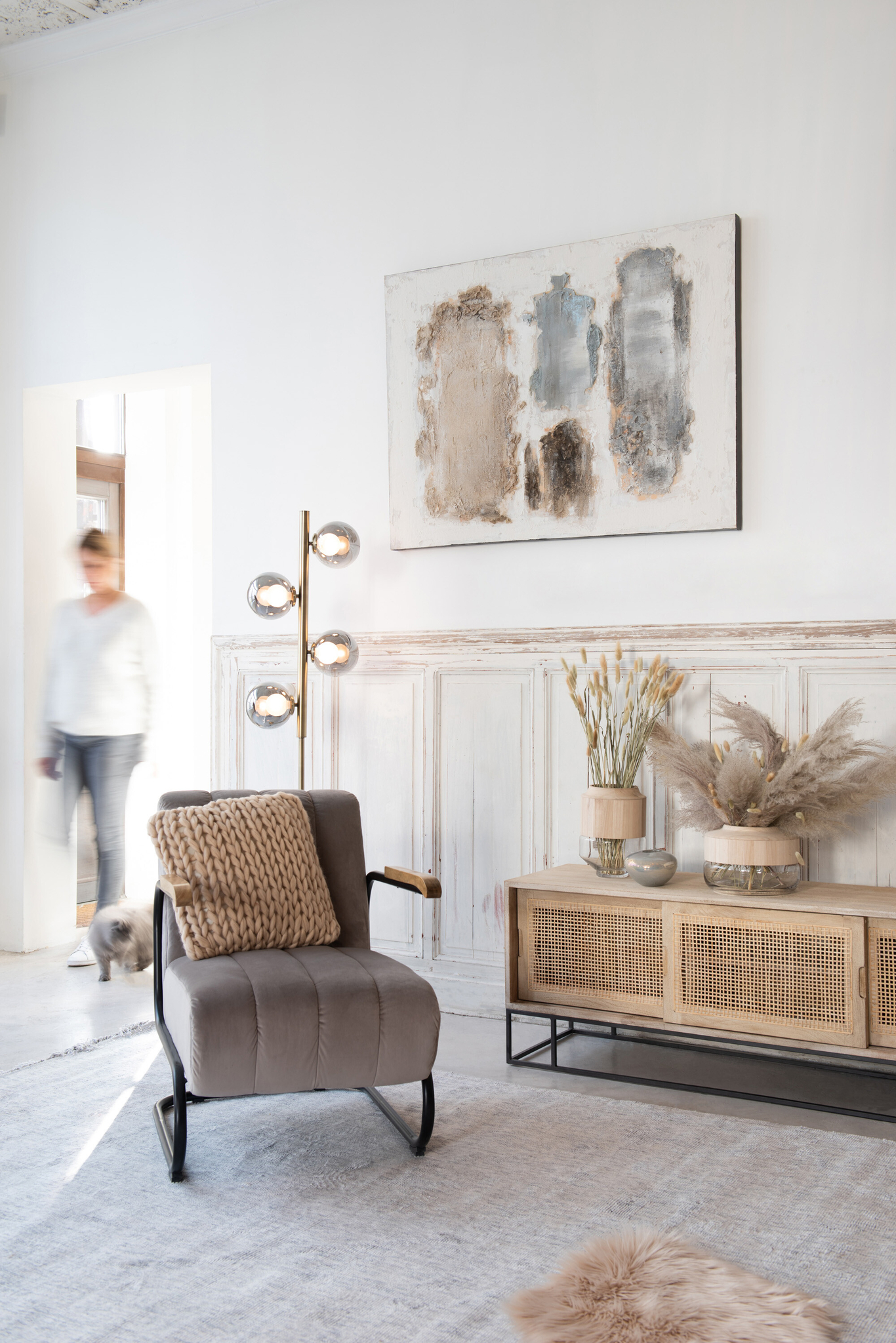 In een lichte, moderne woonkamer staat de Zit Relax 1 Persoon Grijs Textiel fauteuil met een structuurkussen, een houten dressoir met decoratieve vazen, abstracte muurkunst en een gouden vloerlamp. Een persoon en een kleine hond komen binnen door een deuropening op de achtergrond.