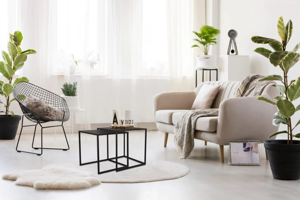 Lichte moderne woonkamer met beige bank, zwarte draadstoel, planten in potten, rond wit vloerkleed, Mirrors and More Bijzettafel JUDY en licht door transparante gordijnen. De inrichting bestaat uit plaids, kussens en minimalistische accessoires.