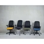 Vier Workliving Nora Mesh Groen Regain ergonomische bureaustoelen (NEN1335), elk met een zwarte netrug en zittingen in geel, zwart, groen en blauw, staan op een rij tegen een lichte muur op een betonnen vloer.