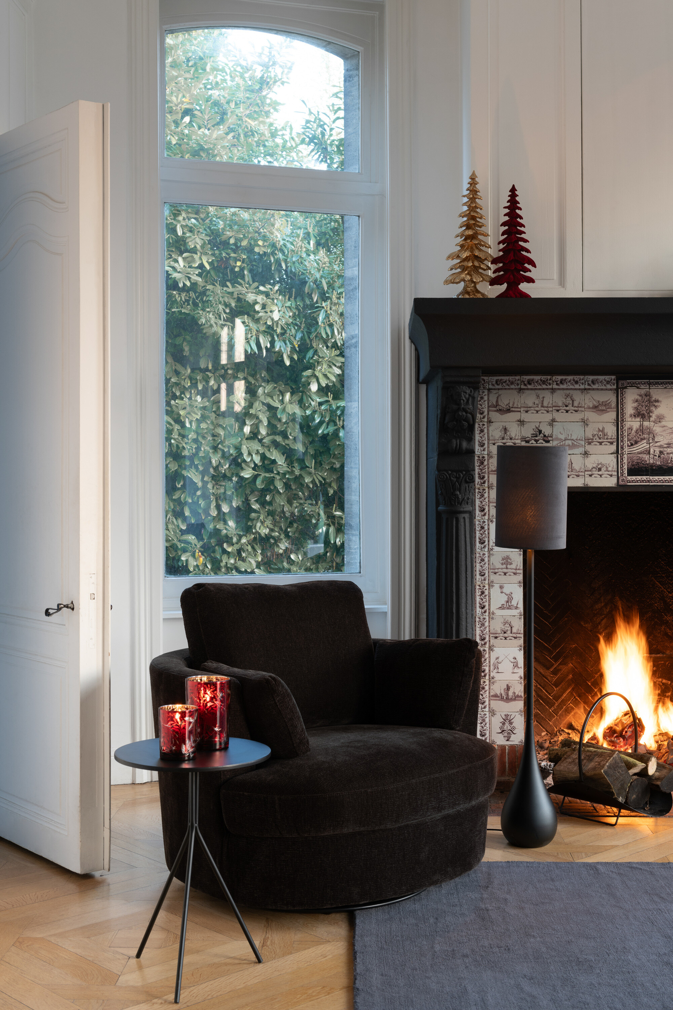 Een gezellige woonkamer met een Draaistoel Populierenhout Donkerbruin, een bijzettafel met rode kaarsen, een brandende open haard en hoge ramen met groen. Op de schoorsteenmantel boven de open haard staan decoratieve rode en gouden boompjes.