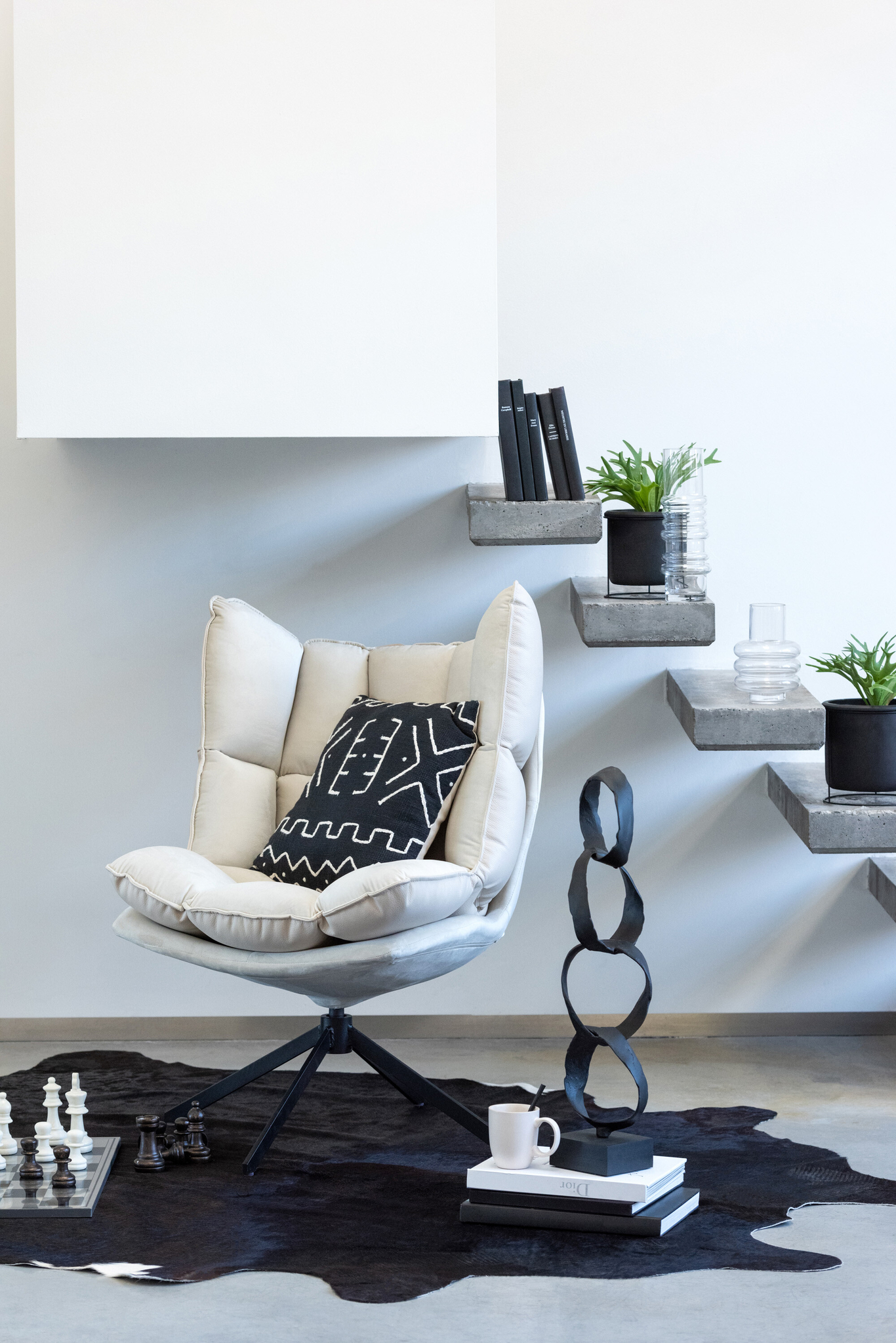 Een moderne leeshoek met de Stoel Relax Kussen Beige Metaal draaistoel, een kussen met patroon, zwart-wit decor, planten in potten op betonnen planken, een kleed van koeienhuid en een schaakbord op de vloer.