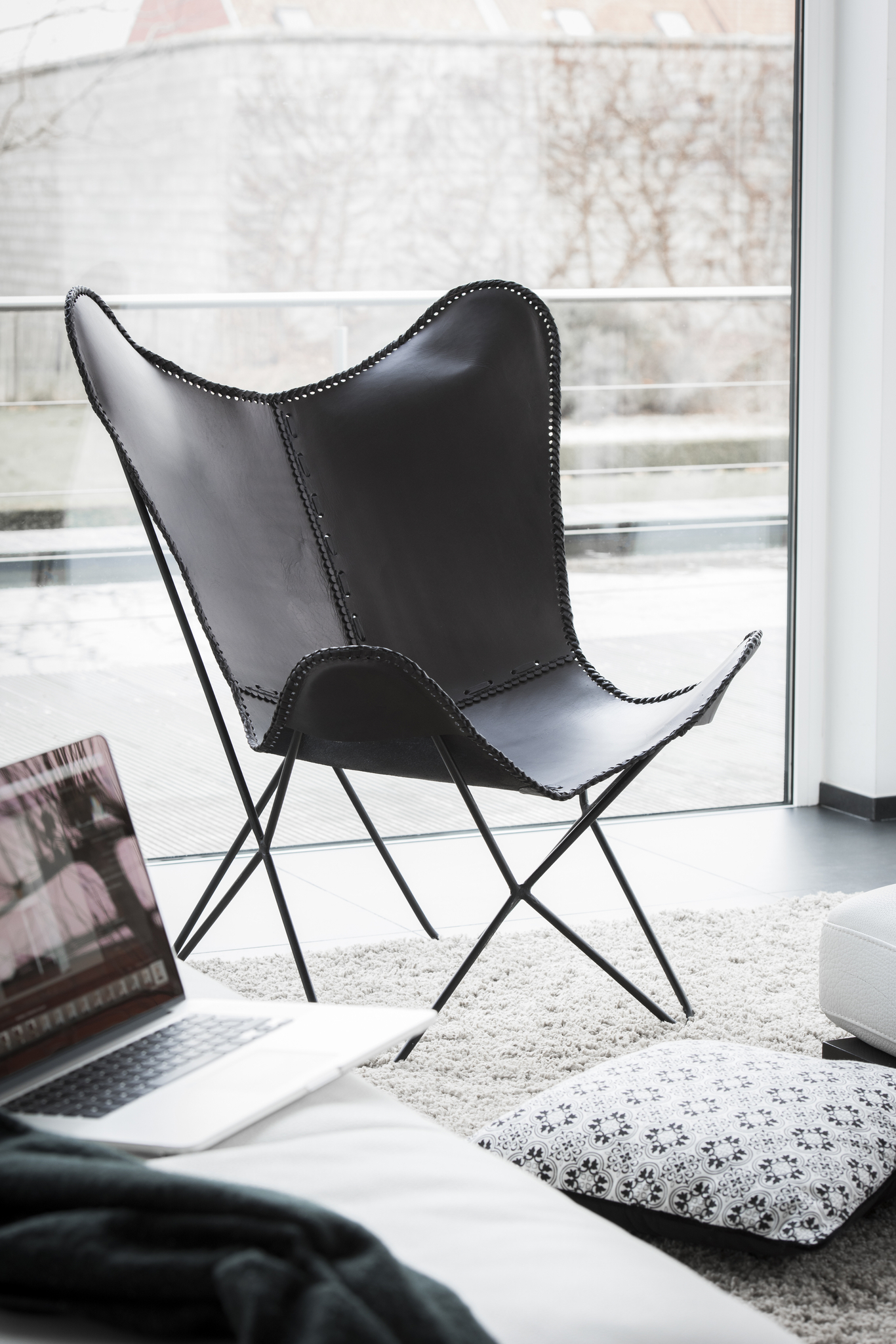 De Lounge Stoel Leer Zwart Metaal, een moderne zwartleren stoel met metalen poten, staat op een licht vloerkleed bij een groot raam. Een kussen met patroon en een open laptop rusten op een witte bank vlakbij, met buiten bomen en gebouwen zichtbaar.