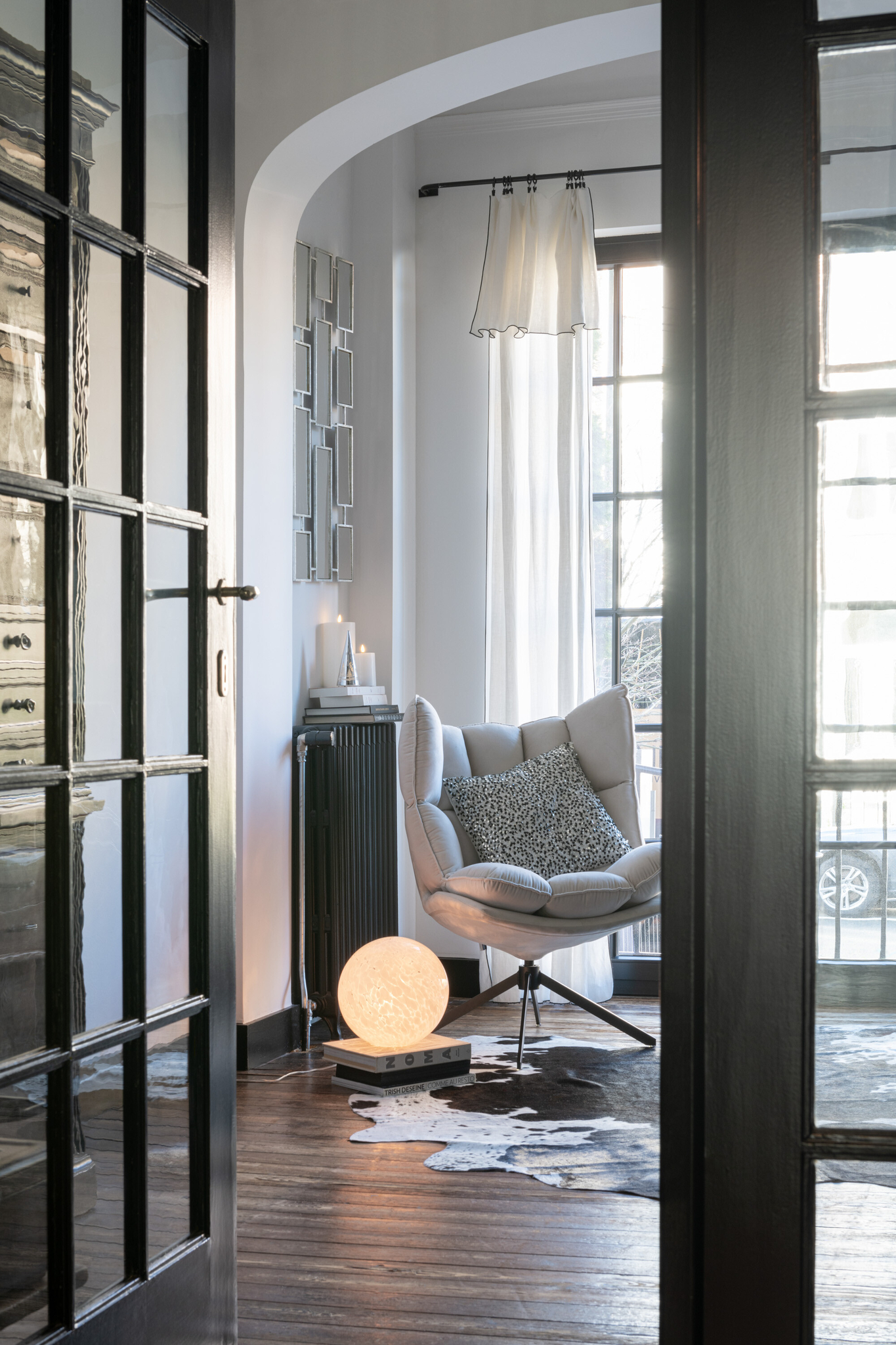 Een gezellige leeshoek met een moderne fauteuil, het Stoel Relax Kussen Extra Lichtgrijs kussen, een gloeiende bollamp op de vloer, een koeienhuid vloerkleed, hoge ramen met transparante gordijnen en houten vloeren bekeken door een deuropening.