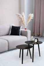 Een moderne woonkamer met een lichtgrijze bank, zwarte kussen, Mirrors and More Bijzettafel VILANA ronde zwarte nesttafels, een wekker, decoratieve beeldjes, een vaas met pampagras en zachtroze gordijnen.