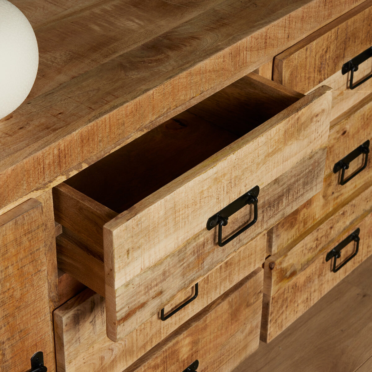 Een close-up van de Dressoir Boaz 200 cm, een houten kast met verschillende lades, waarvan één open, met een natuurlijke rustieke afwerking en zwarte metalen handgrepen. Linksboven is een deel van een wit rond object zichtbaar.