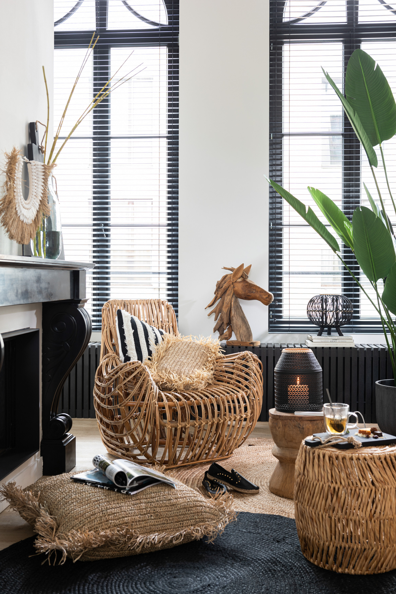 Een gezellige woonkamer is voorzien van de Stoel Ana Rotan Natuurlijk fauteuil, een gestreept kussen, geweven voetenbank, jute vloerkleed, potplant, theeset, tijdschrift, decoratieve paardensculptuur en grote ramen met zwarte jaloezieën.