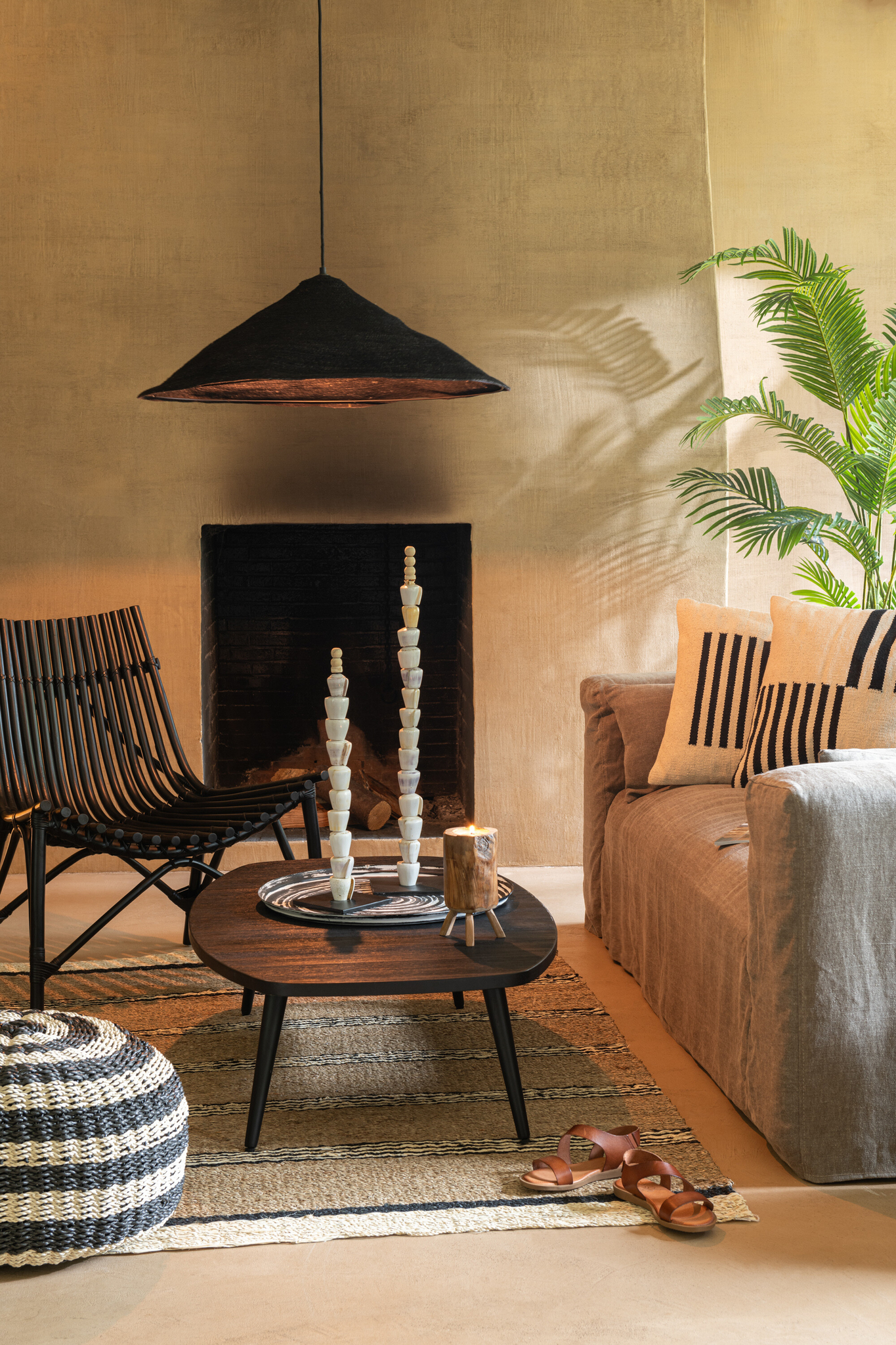 Een gezellige woonkamer is voorzien van de Stoel Rotan Zwart Handgemaakt, een grijze bank met gestreepte kussens, een ronde houten salontafel, gestreepte poef, geweven vloerkleed, decoratieve kandelaars, palm in pot, sandalen op de vloer en een hanglamp.