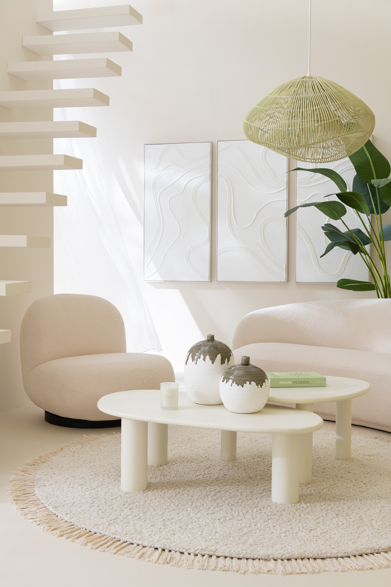 Een moderne, minimalistische woonkamer met gebroken witte loungestoel Sneeuw Wit/Zwart Comfort, een rond vloerkleed, abstracte muurkunst, zwevende trap, grote plant, geweven hanglamp en twee vazen met boeken op een ovale salontafel.