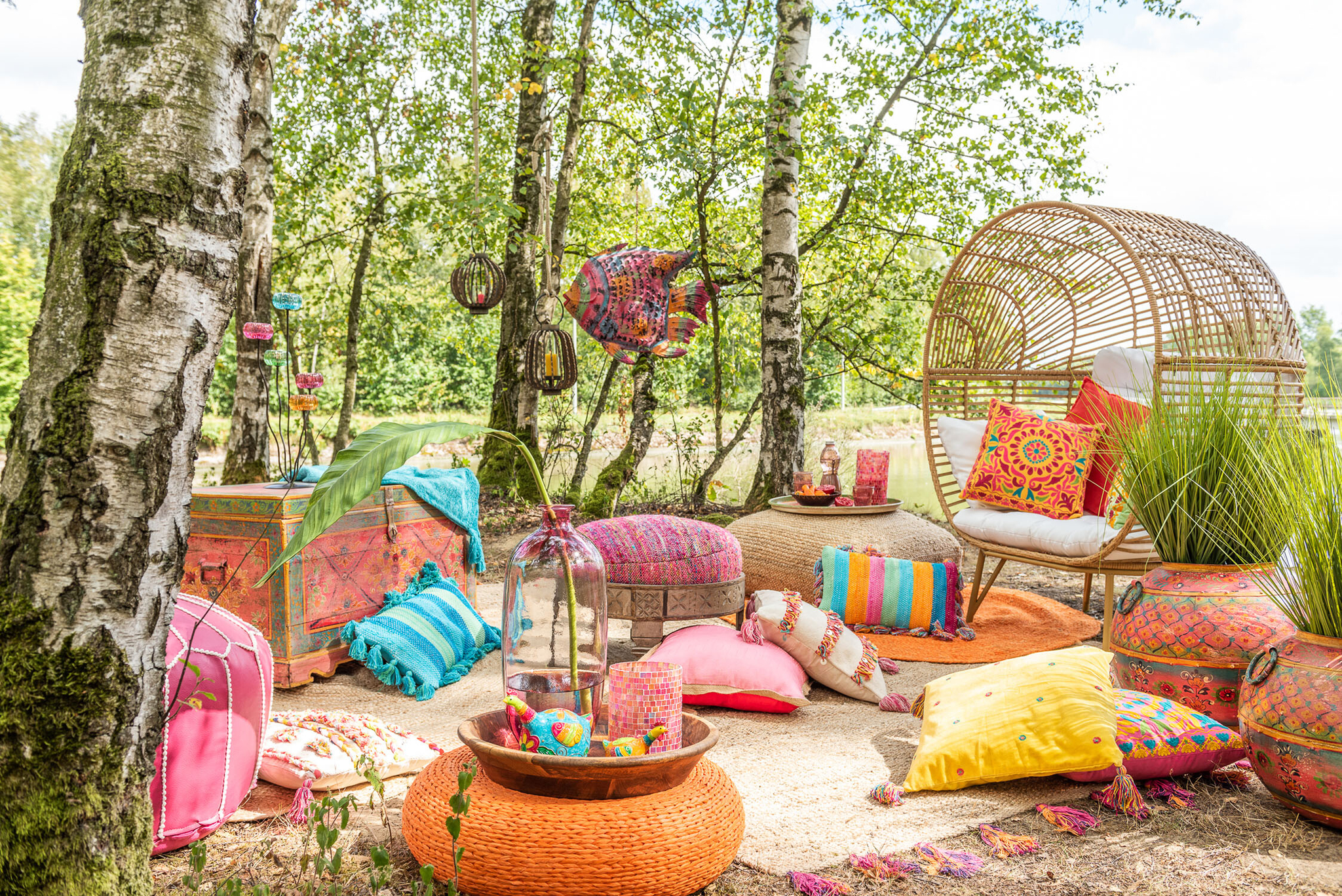 Een gezellige zithoek buiten met de Lounge Stoel Ovaal Natuurlijk Staal, heldere bohemien inrichting, kleurrijke kussens en kleden en lantaarns. Levendig roze, oranje en gele accenten creëren een vrolijke sfeer tussen de berkenbomen.