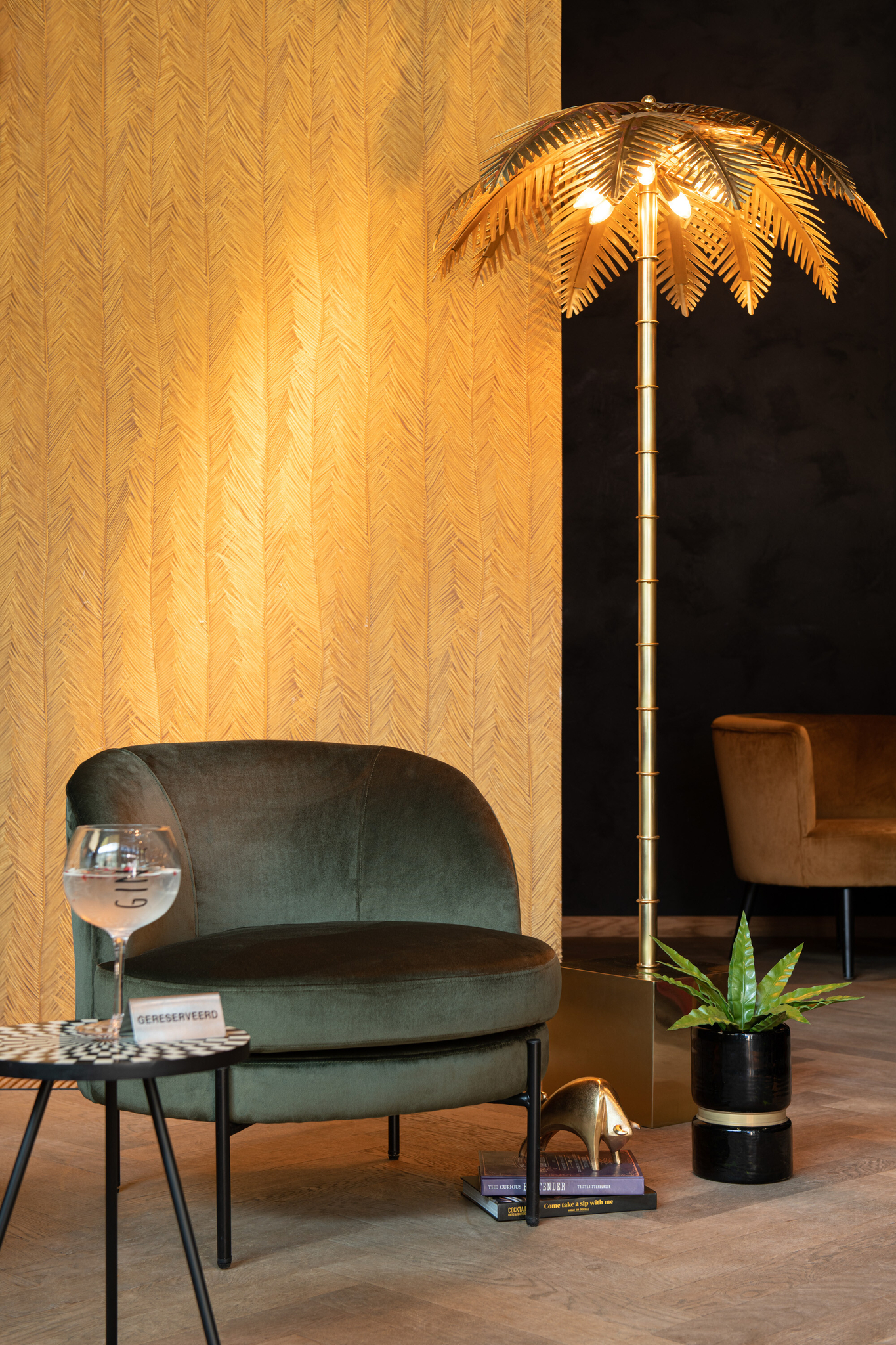Een moderne lounge is voorzien van de Lounge Stoel Rond Groen Textiel, een tafeltje met een drankje en boeken, een gouden vloerlamp met palmboom, een potplant en een muur met bruin patroon en warme verlichting.