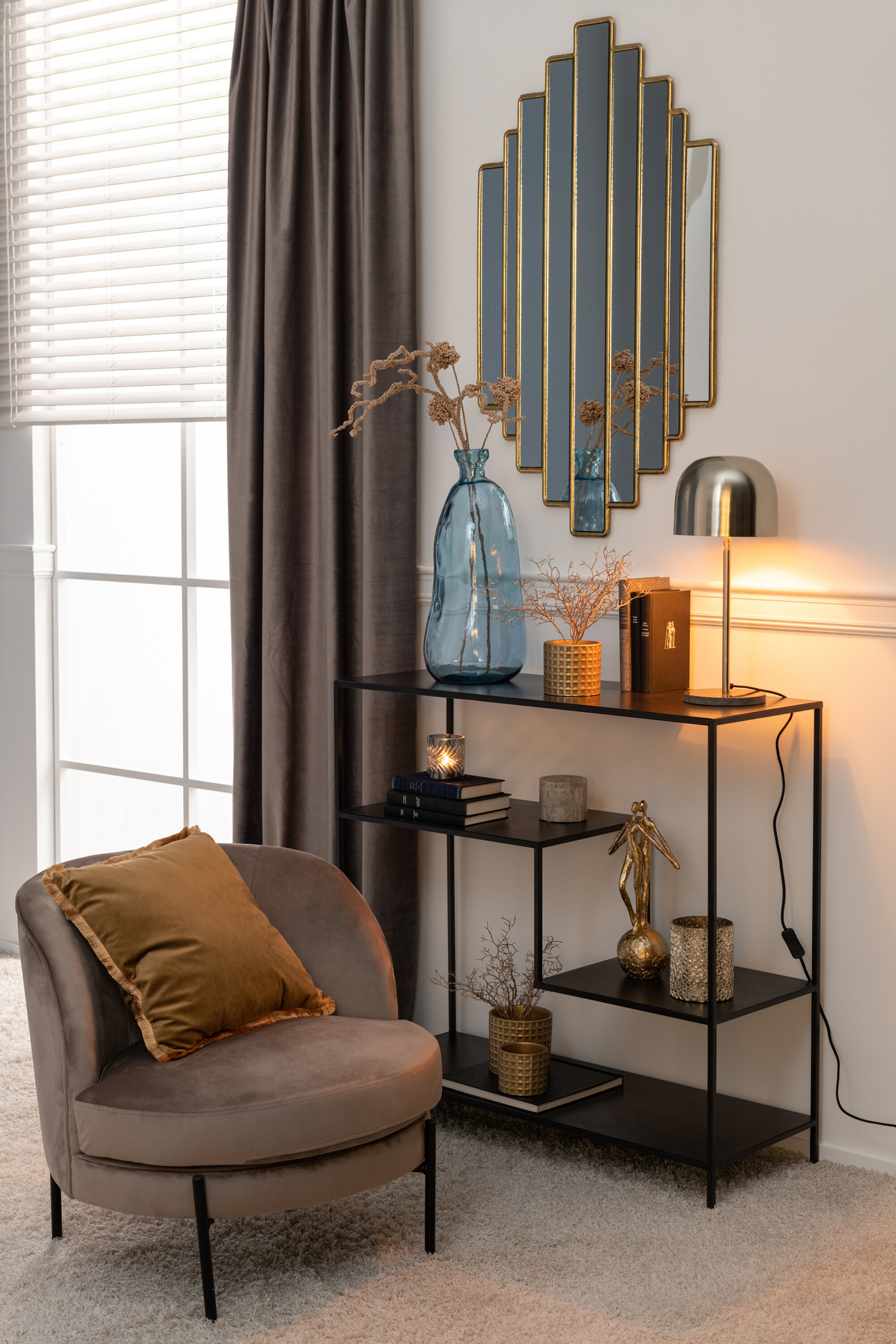 Een knus hoekje met de Lounge Stoel Rond Bruin Textiel en een gouden kussen staat naast een zwarte metalen plank met boeken, decor en een blauwe vaas met gedroogde bloemen. Boven hangen een gouden lamp en een art-decospiegel aan de crèmekleurige muur.