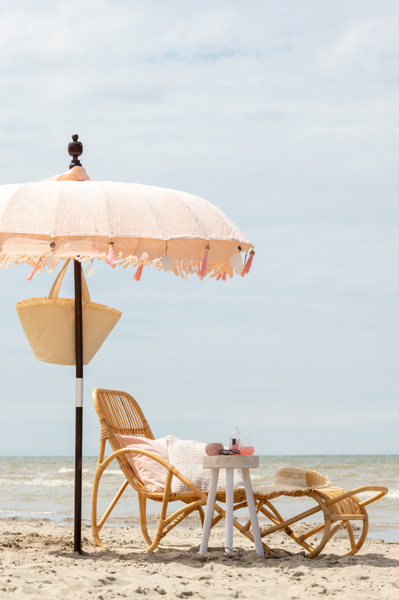 De Ligstoel Els Rotan Natuurlijk met een roze kussen rust onder een parasol met franjes op het zandstrand, naast een tafel met drankjes en een zonnehoed, met de oceaan en de lucht op de achtergrond.