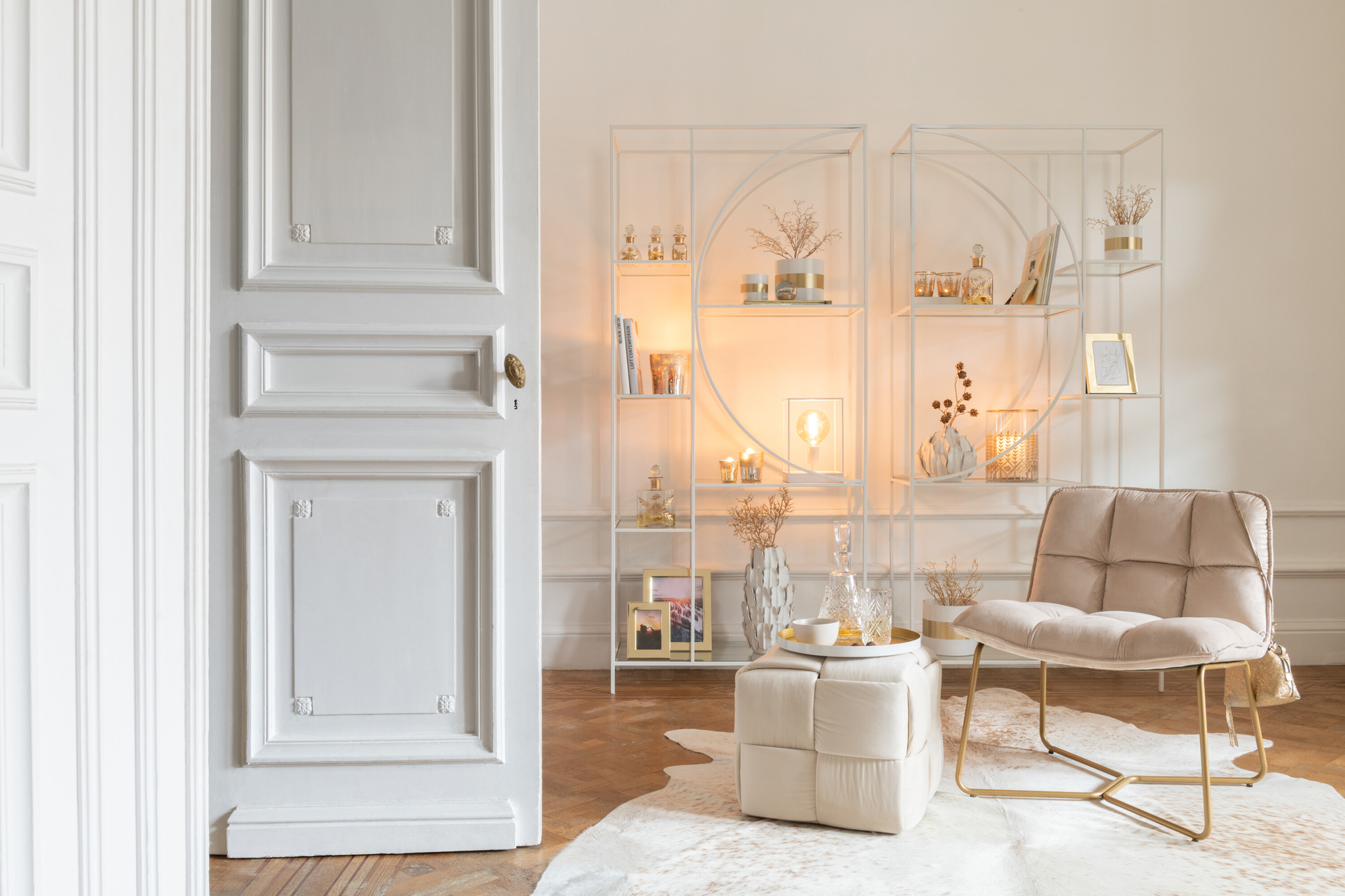 Een gezellige, moderne woonkamer is voorzien van de Ligstoel Lisa Beige Metaal/Textiel fauteuil met voetenbank, een wit koeienhuid vloerkleed, minimalistische gouden planken met decor en planten, zachte verlichting en een open witte deur op de voorgrond.