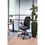 Een moderne kantoorruimte met de Workliving Nora Mesh Blauw Regain ergonomische stoel (NEN1335), een staand bureau met een lamp en mokken, en een hoge groene plant in een grote pot, tegen matglazen panelen.