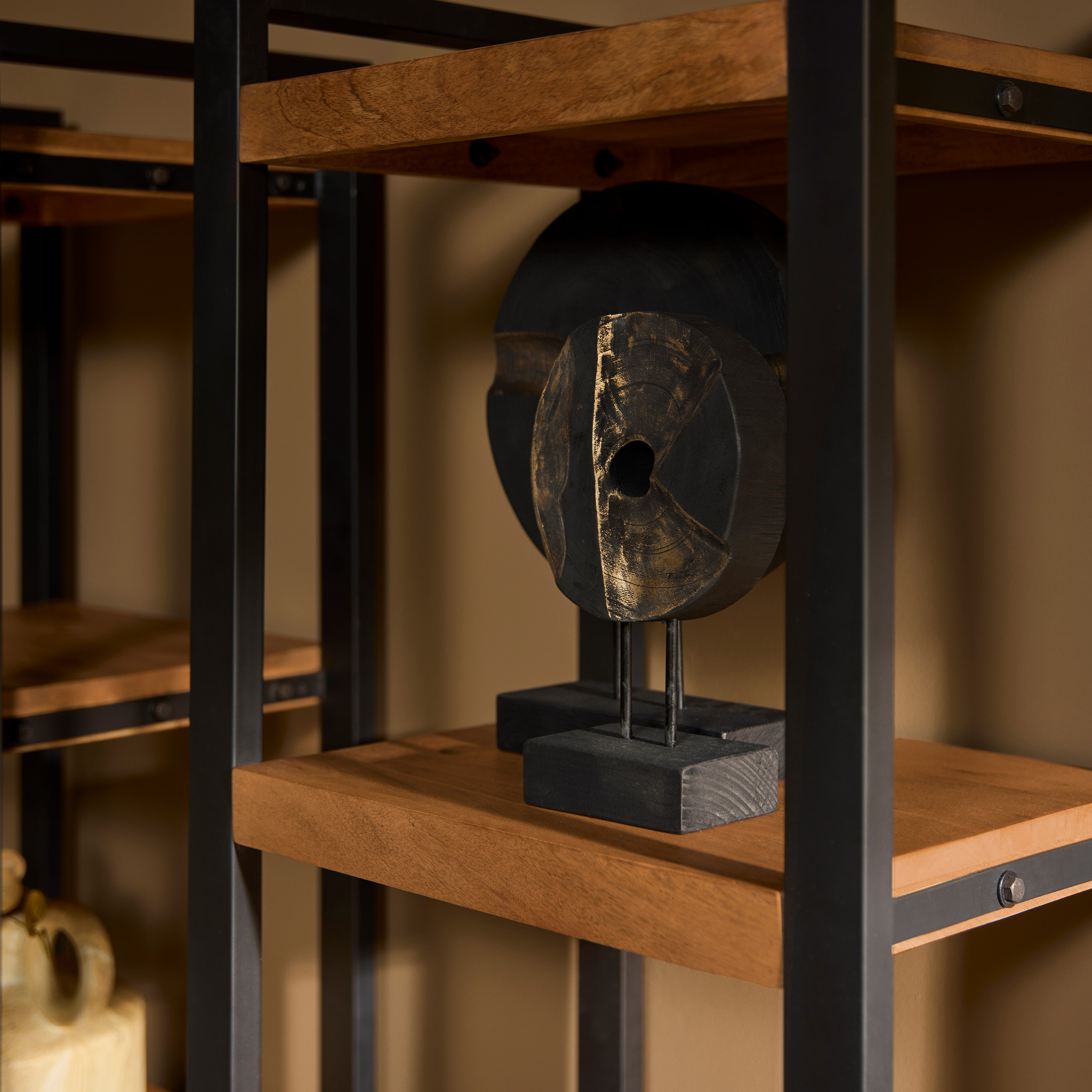 Een close-up van een decoratieve zwart-gouden abstracte sculptuur op een Boekenkast Boaz | 1 deur houten plank met metalen frame tegen een beige muur, met andere objecten en planken op de achtergrond.