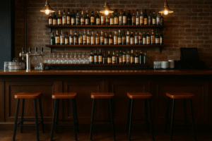 Een gezellige gemiddelde bar met vier krukken, een houten toonbank, planken met flessen drank, wijnglazen, gestapelde borden en drie hangende hanglampen tegen een bakstenen muur.