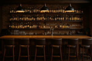 Een gezellige, gemiddelde bar met vijf bruine leren krukken, een houten toonbank, planken vol drankflessen en glaswerk en drie hanglampen tegen een zichtbare bakstenen muur.