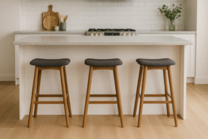 Een moderne keuken met een wit eiland, drie houten krukjes met donkere kussens, een lichthouten vloer en een witte tegelachterwand. Keukengerei en een plant staan op het aanrecht op de achtergrond - hoeveel ruimte heb je nodig voor jouw droomopstelling?.