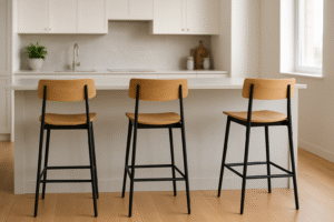 Drie houten barkrukken met zwarte metalen poten staan op een rij bij een wit keukeneiland, en laten duidelijk zien hoeveel ruimte je nodig hebt tussen elke stoel in deze lichte, moderne keuken met lichte houten vloeren en witte kasten.