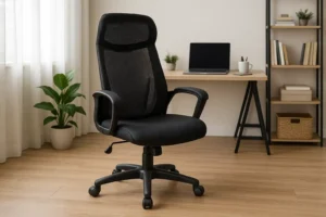Een zwarte ergonomische bureaustoel, perfect voor wie let bij een goede bureaustoel, staat op een houten vloer in een thuiskantoor. Een bureau, laptop, planten, boeken en planken completeren de ruimte die baadt in het natuurlijke licht van een raam met witte gordijnen.