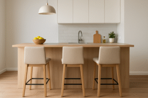 Een moderne, minimalistische keuken met een licht houten eiland op de ideale hoogte voor een bar, drie beige barkrukken, een schaal citroenen, hanglamp, witte kasten, gootsteen en eenvoudige decoratie zoals een snijplank en een klein plantje.