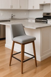 Een moderne keuken met een licht houten vloer, witte kasten en een granieten aanrechtblad. Een grijs gestoffeerde aanrechtstoel met houten poten en een metalen voetsteun staat voor het kookeiland.