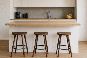 Een moderne keuken heeft een eiland met een licht houten aanrechtblad en drie houten barkrukken op goede barhoogte, een gootsteen, een koffiezetapparaat en schalen met bananen tegen een neutrale grijze achterwand. Witte kasten dragen bij aan de strakke, minimalistische look.