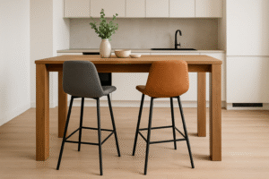 Een moderne keuken met een houten tafel aan zithoogte bij bartafel, twee stoelen - een grijze en een bruine - zwarte metalen poten, een vaas met groene bladeren en een kom. Witte kastjes en een zwarte kraan staan op de achtergrond.